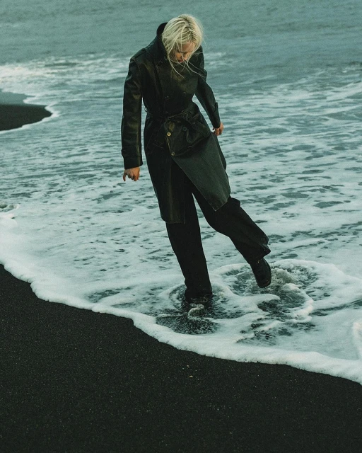 Woman in a black trench coat walks through the surf on a black sand beach with ocean waves, creating a fashion editorial image.