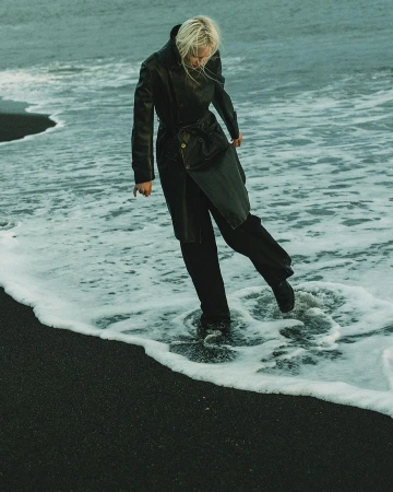 Woman in a black trench coat walks through the surf on a black sand beach with ocean waves, creating a fashion editorial image.