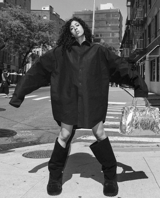 A woman poses in a city street wearing an oversized black button-down shirt and boots, holding a silver handbag in black and white.