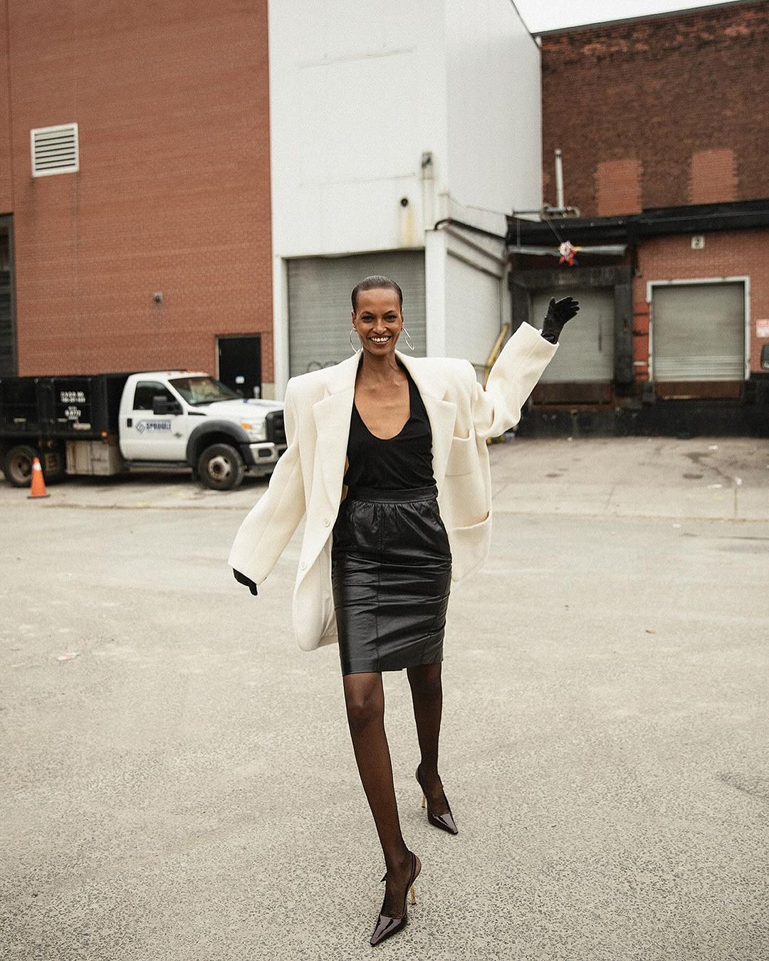 Elegant Black woman in a beige blazer and black skirt strikes a pose on an urban street, with a white truck in the background.