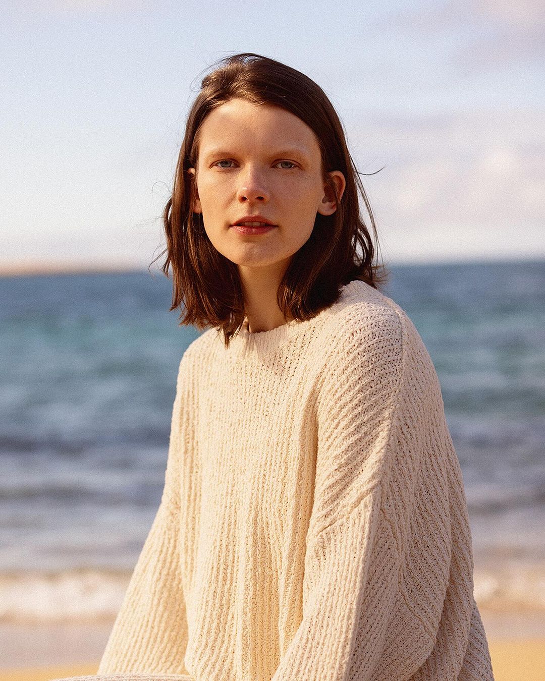 A woman in a knitted sweater poses on a sandy beach with a blurred ocean background under a bright sky in a medium outdoor shot.