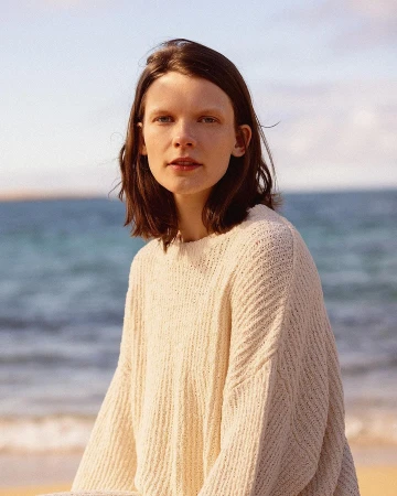A woman in a knitted sweater poses on a sandy beach with a blurred ocean background under a bright sky in a medium outdoor shot.
