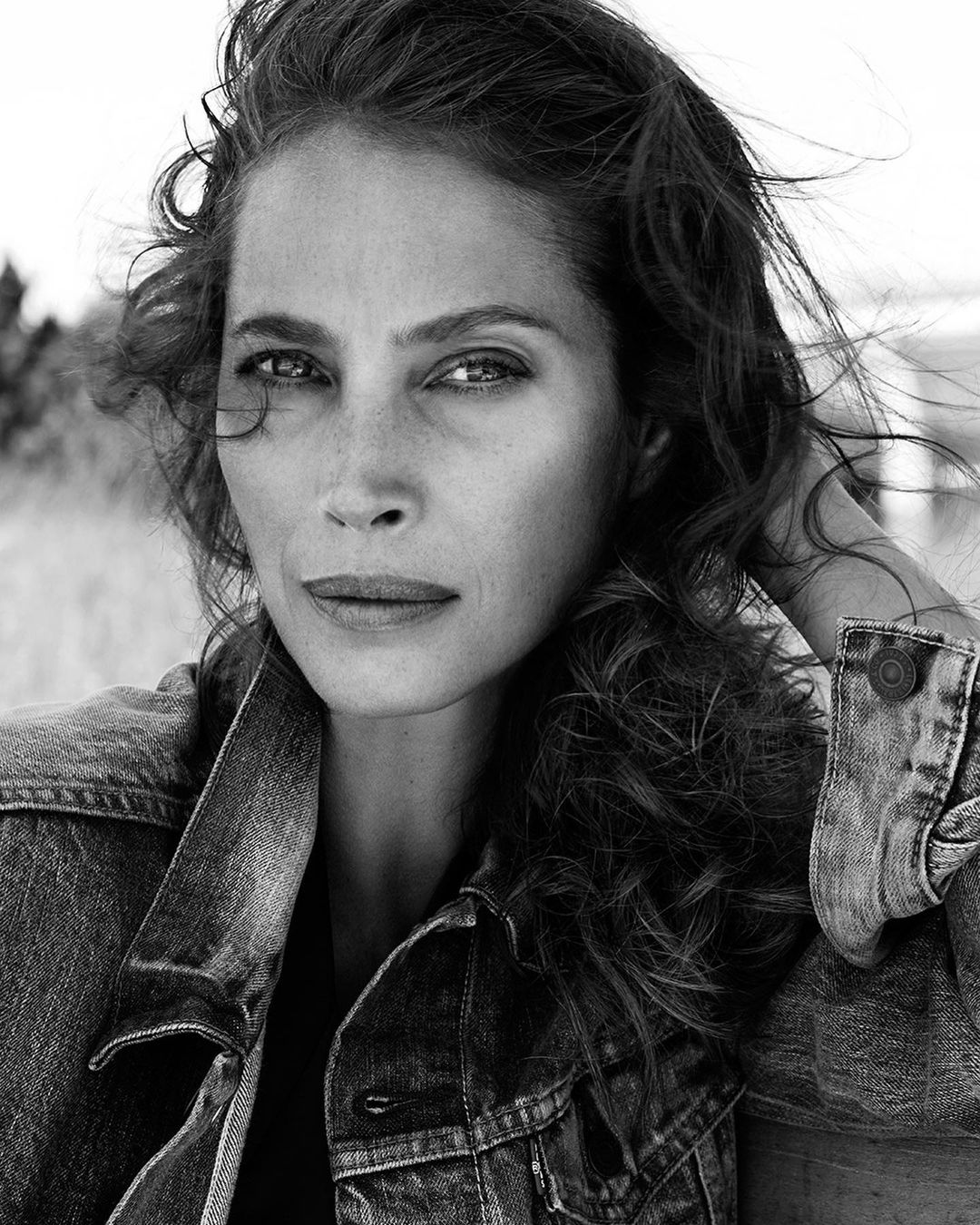 Black and white portrait of an adult woman with wavy hair wearing a denim jacket, posing outdoors with a neutral facial expression.