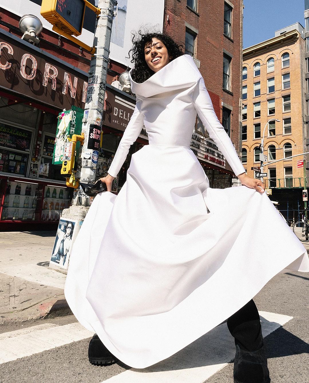 A smiling woman in a billowing white dress stands on a crosswalk in a city street with buildings and signage visible in the background.