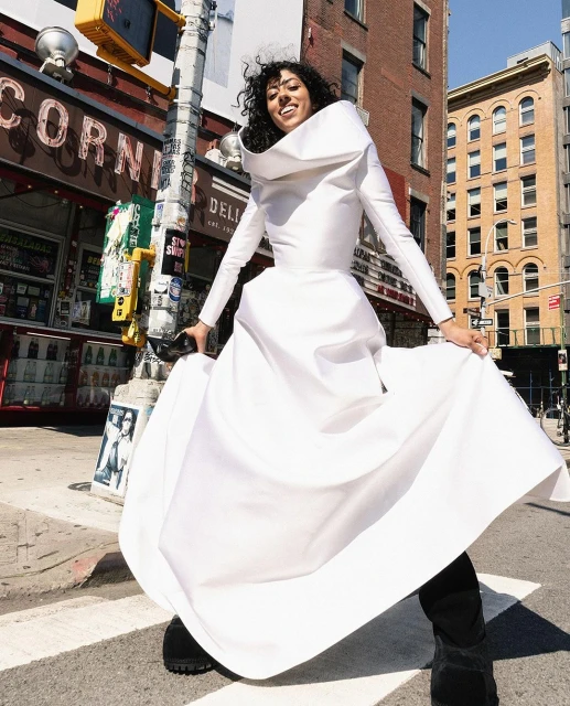 A smiling woman in a billowing white dress stands on a crosswalk in a city street with buildings and signage visible in the background.