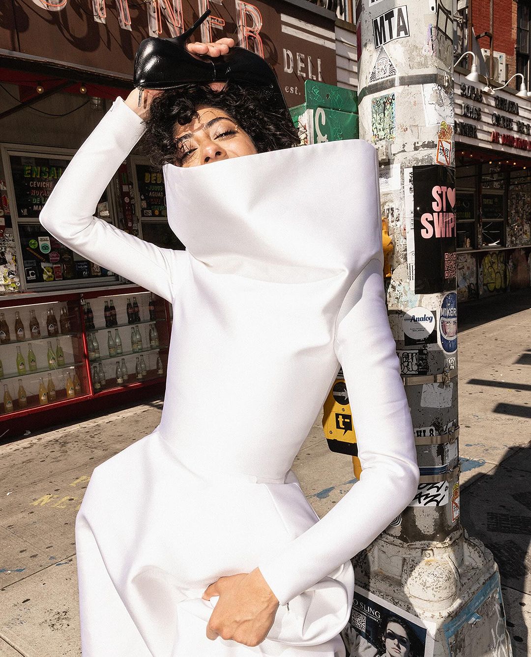 Woman in an oversized white gown posing with black high heel shoes in an urban setting with a deli and theater in the background.