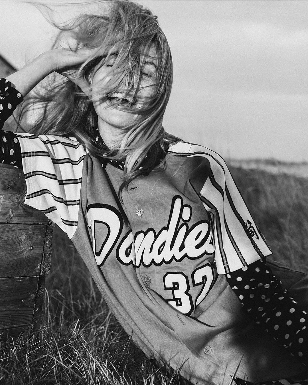 Monochrome portrait of a smiling woman in a baseball jersey outdoors with windblown blonde hair and grassy background, leaning against a wooden structure.