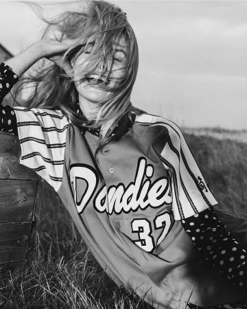 Monochrome portrait of a smiling woman in a baseball jersey outdoors with windblown blonde hair and grassy background, leaning against a wooden structure.