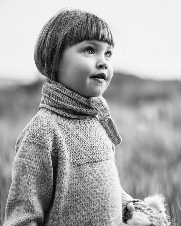 Black and white portrait of a child with short bob hair wearing a knitted patterned sweater outdoors.
