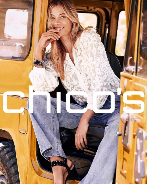 A smiling adult woman is sitting inside a yellow vehicle, wearing jeans and a white patterned shirt. The brand name CHICO'S is visible.