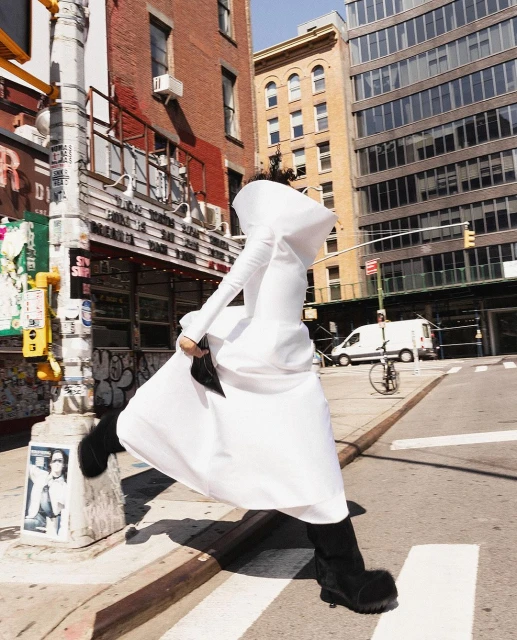A model in a white hooded dress walks across a crosswalk in a city setting with buildings and a street light in the background.