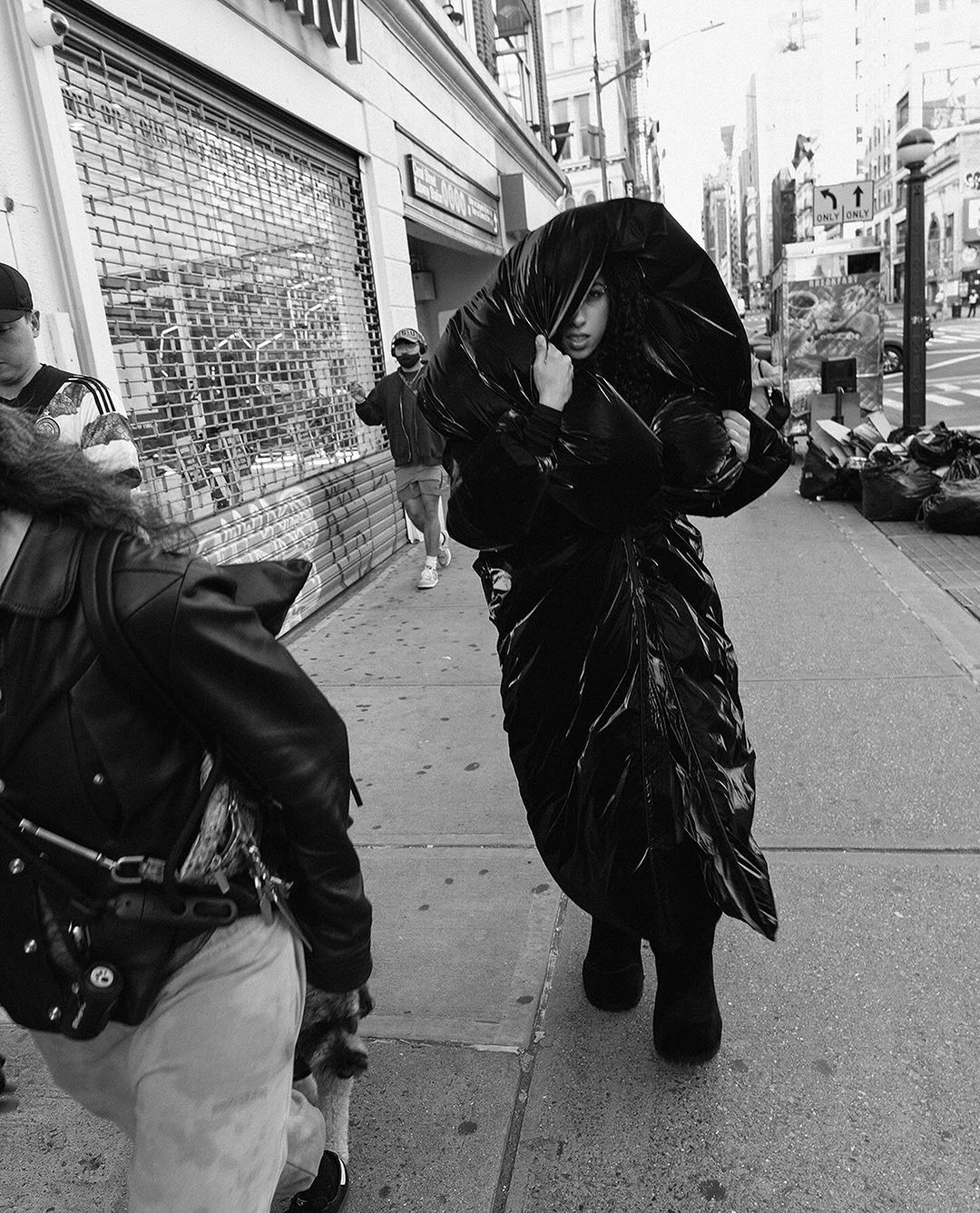 Monochrome shot of a woman walking on a city sidewalk wearing an avant-garde, voluminous black garment, with other pedestrians nearby.