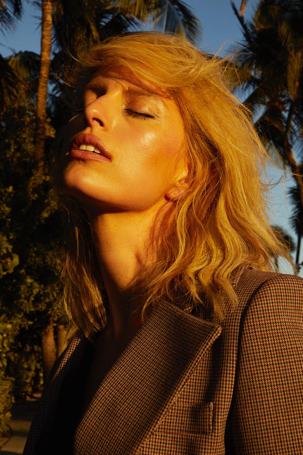 Close-up of a fair-skinned blonde woman in a blazer, head tilted back, eyes closed, bathed in golden sunlight, with palm trees in background.