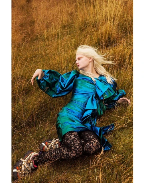 Fashion shot of a woman in a bright turquoise dress with flowing sleeves, lying in a field of golden grass, wearing floral lace tights.