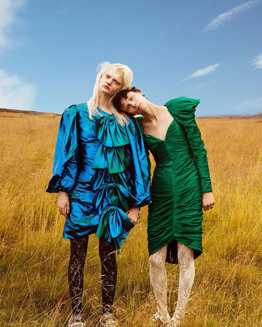 Two fashion models pose in a grassy field under a blue sky. The women wear vintage-style dresses and pose together in a landscape. 