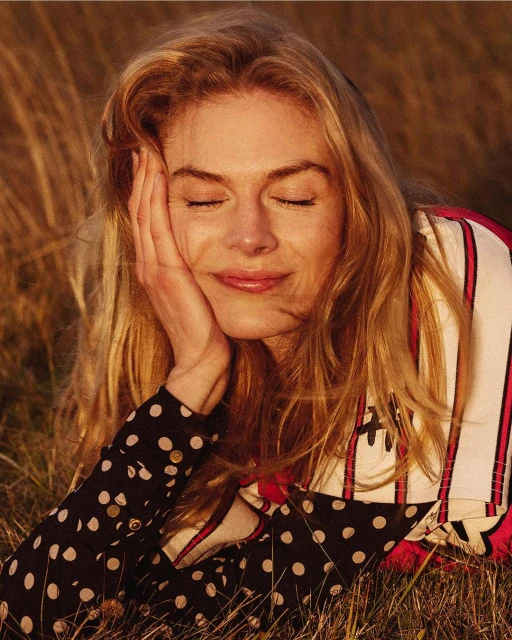 Blond woman with eyes closed lying in tall grass, wearing polka dot top, bathed in golden sunlight, creating a serene, peaceful portrait.