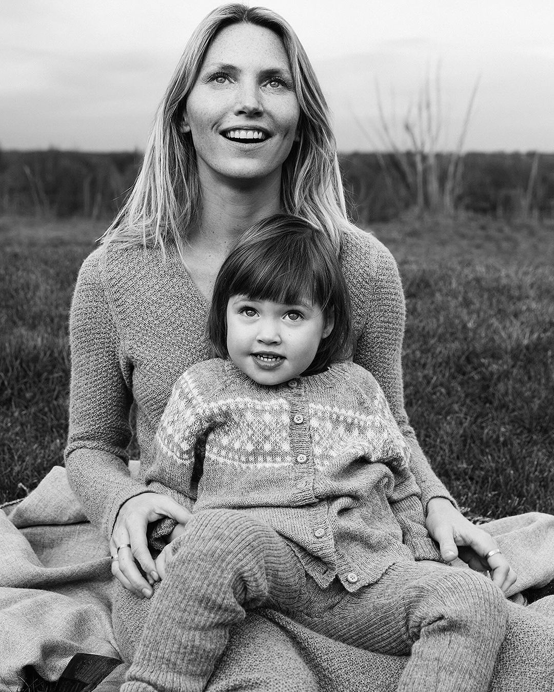 Black and white portrait of a blonde mother and young child in matching knitwear, seated outside on a blanket in a grassy field.