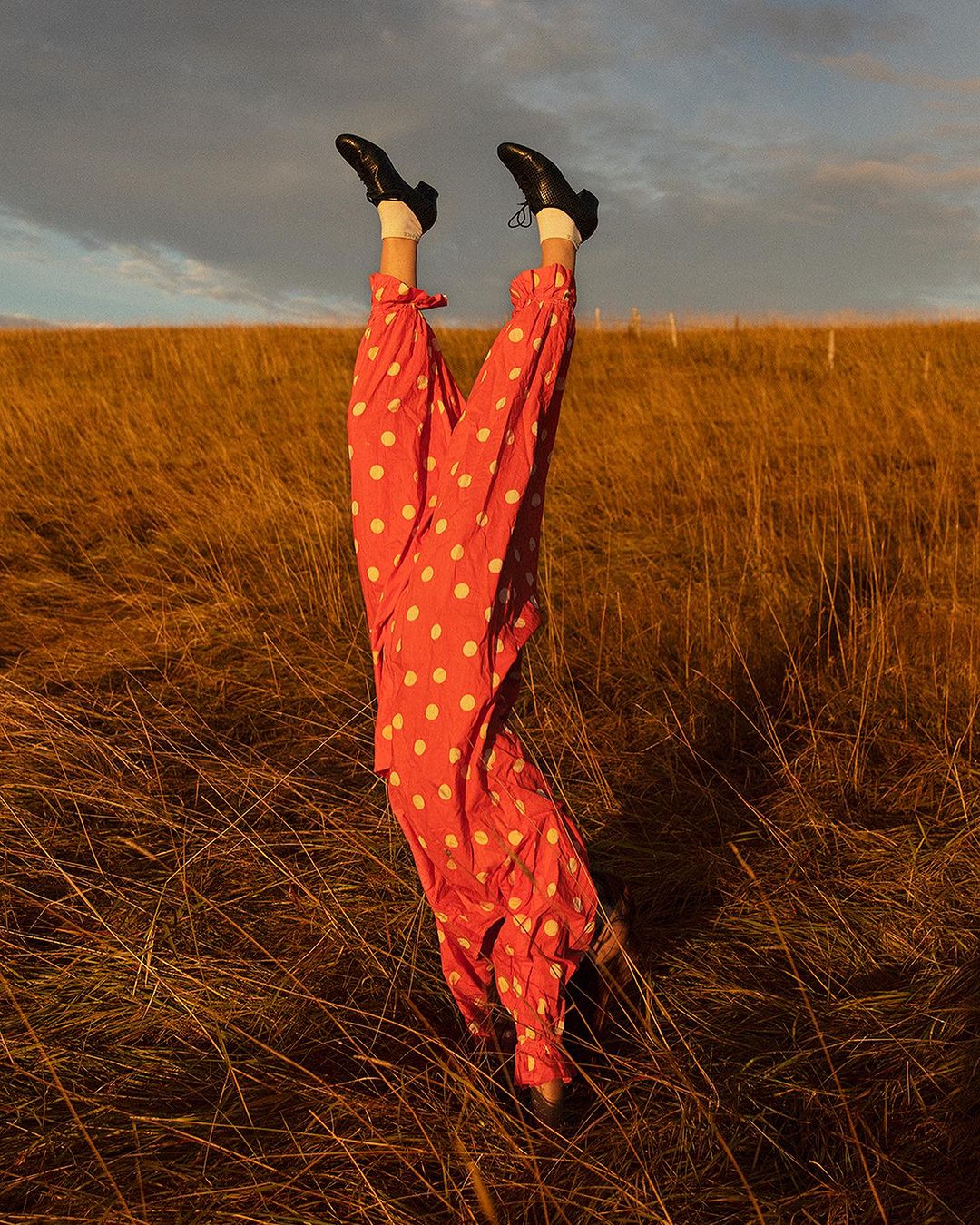 Person in red polka dot pants and black shoes does a handstand in a field of tall, golden grass under a cloudy sky.