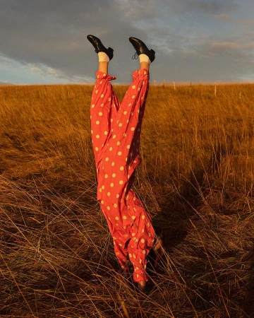 Person in red polka dot pants and black shoes does a handstand in a field of tall, golden grass under a cloudy sky.