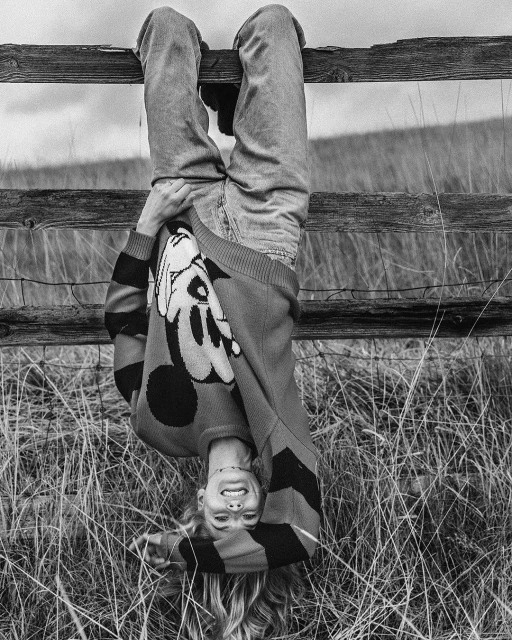 Black and white photo of a smiling adult female hanging upside down on a wooden fence in a rural field wearing a cartoon sweater.