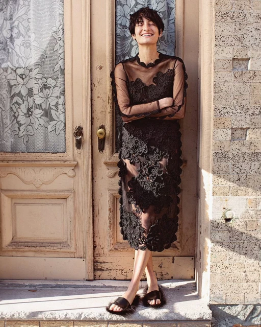 A woman with short hair poses confidently in a black lace dress against a textured door, creating a striking fashion portrait.