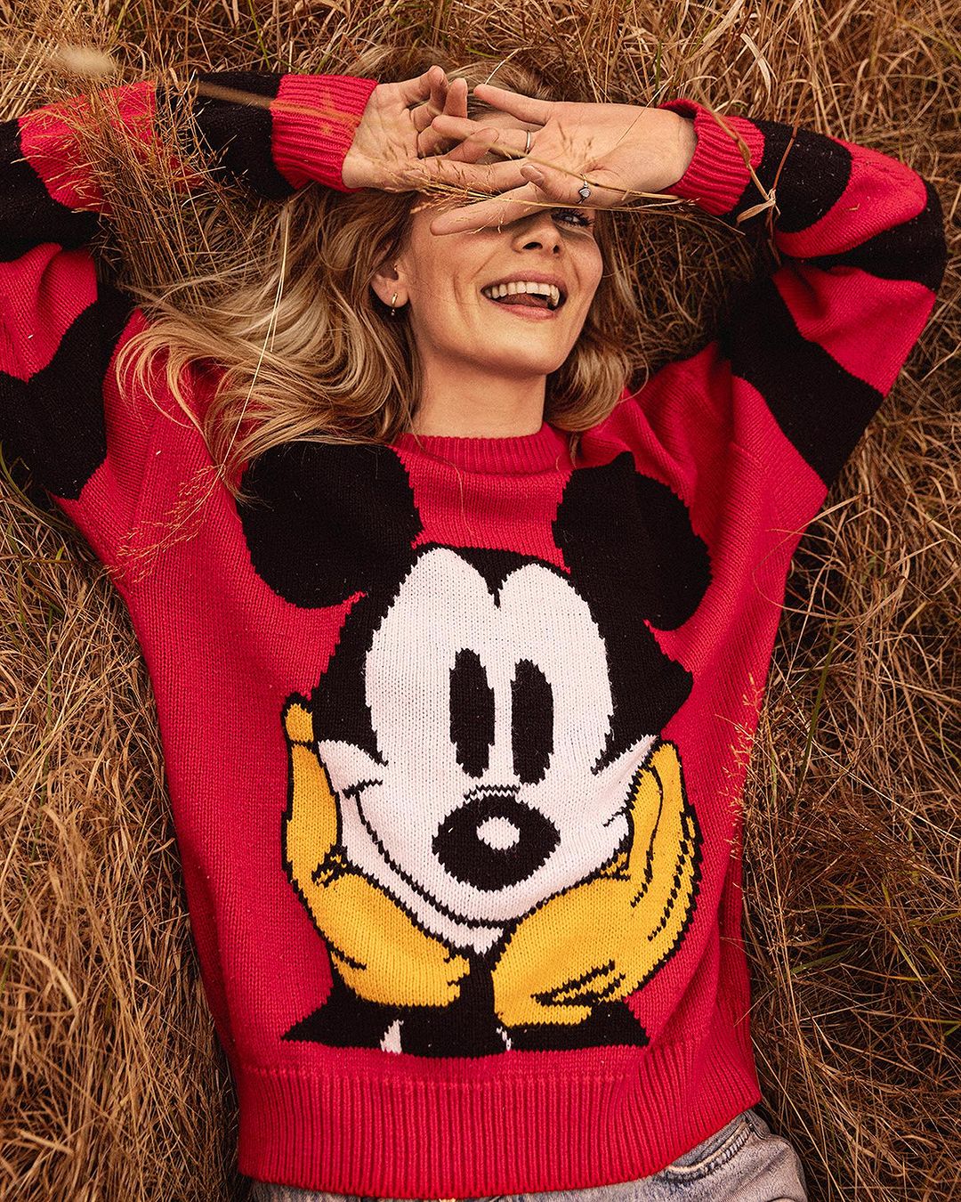 Blonde woman in Mickey Mouse sweater lying in field of dried grasses, smiling, hands over face, low angle, casual fashion.