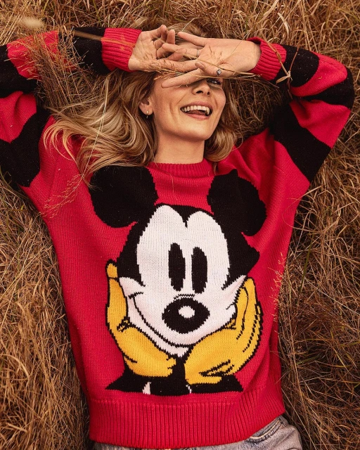 Blonde woman in Mickey Mouse sweater lying in field of dried grasses, smiling, hands over face, low angle, casual fashion.