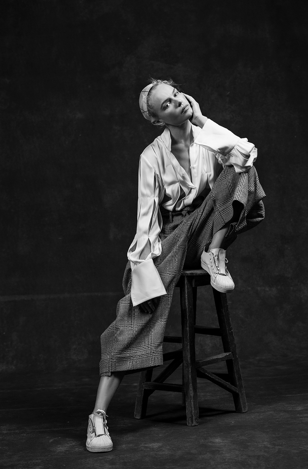 Blonde adult female fashion model with hairband, wearing white blouse and plaid pants, seated on wooden stool against dark studio backdrop.