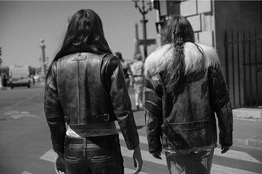 Two people in leather jackets walk away on a city street in a black and white photograph taken during the day. Fashion and city life.