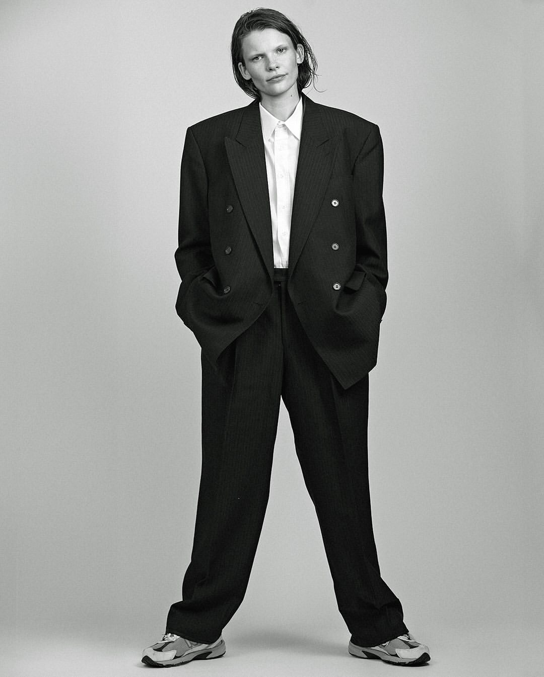 A person with short hair stands in a photo studio wearing a pinstripe suit, white shirt, and athletic shoes, captured in black and white.
