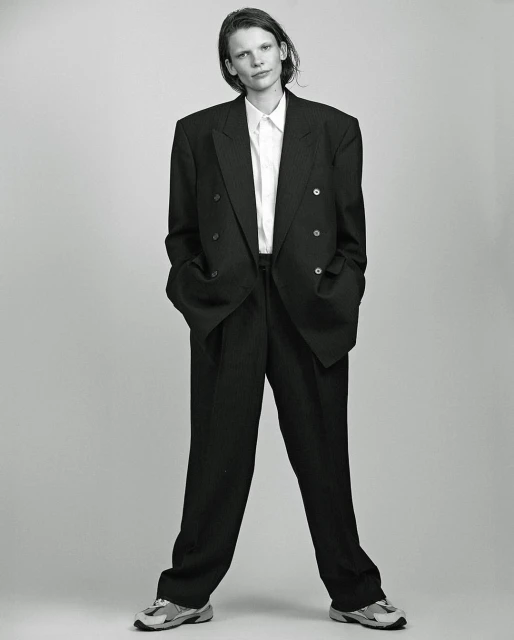 A person with short hair stands in a photo studio wearing a pinstripe suit, white shirt, and athletic shoes, captured in black and white.