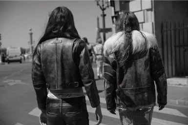 Two women with long hair wearing leather jackets walking down a city street viewed from behind in black and white.