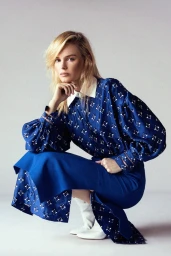 Blonde female model in patterned blue dress and white boots squatting against a plain white studio background