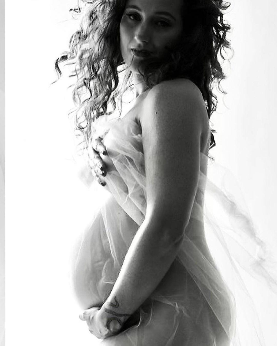 Black and white portrait of a pregnant woman partially covered with sheer white fabric, posing in a brightly lit indoor environment.