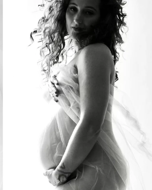 Black and white portrait of a pregnant woman partially covered with sheer white fabric, posing in a brightly lit indoor environment.