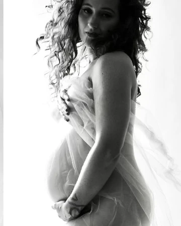 Black and white portrait of a pregnant woman partially covered with sheer white fabric, posing in a brightly lit indoor environment.