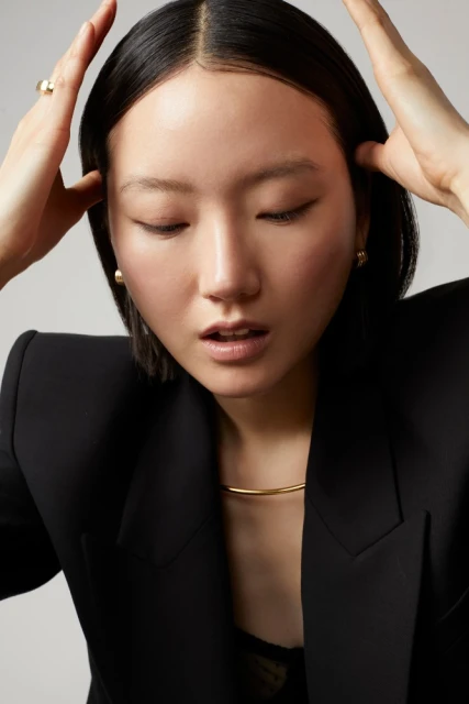 Portrait of an adult woman with her hands near her temples, wearing a black blazer and gold choker necklace, set against a grey backdrop.