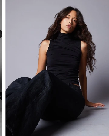 A seated woman wearing a black sleeveless turtleneck and dark trousers against a plain grey studio backdrop.