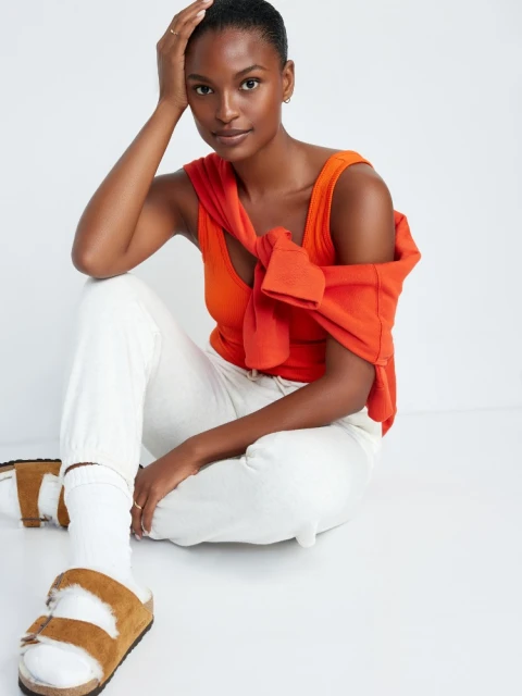 Woman in orange top and white pants sits on floor with sweater around shoulders and casual shoes. Relaxed pose in studio.