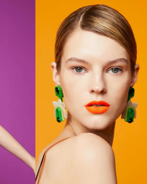 Headshot of fair-skinned model with orange lipstick and large green and white flower earrings against an orange and purple backdrop.