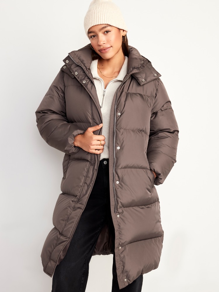 Adult female wearing a long, brown puffer coat and cream-colored beanie in a bright, neutral photo studio setting.