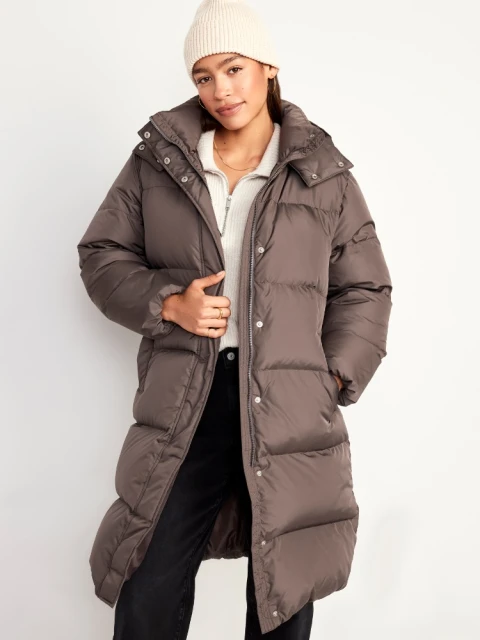 Adult female wearing a long, brown puffer coat and cream-colored beanie in a bright, neutral photo studio setting.