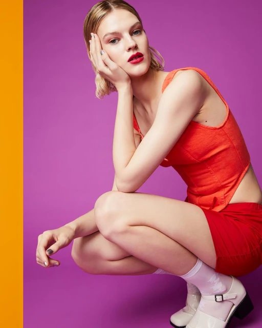 A fair-skinned adult woman in a red outfit poses crouching against a purple background with a vertical orange block on the left side.