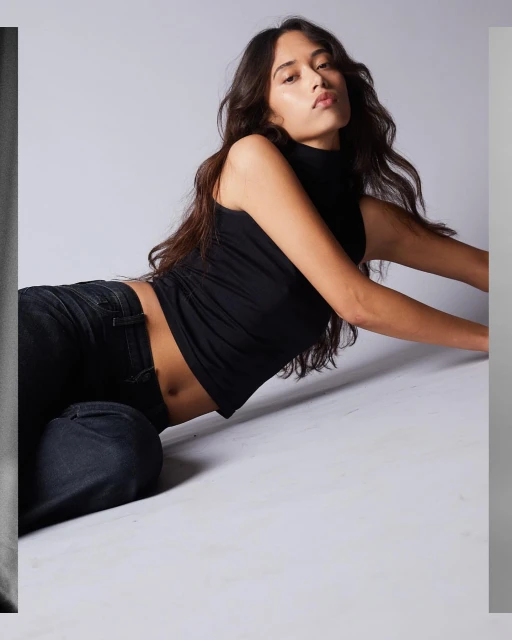 A woman poses in a black tank top and jeans, lying on a grey studio backdrop, her dark hair cascading around her.