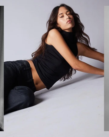 A woman poses in a black tank top and jeans, lying on a grey studio backdrop, her dark hair cascading around her.