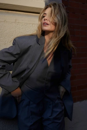 Blonde woman wearing a grey blazer and vest standing outdoors against a brick and stone wall