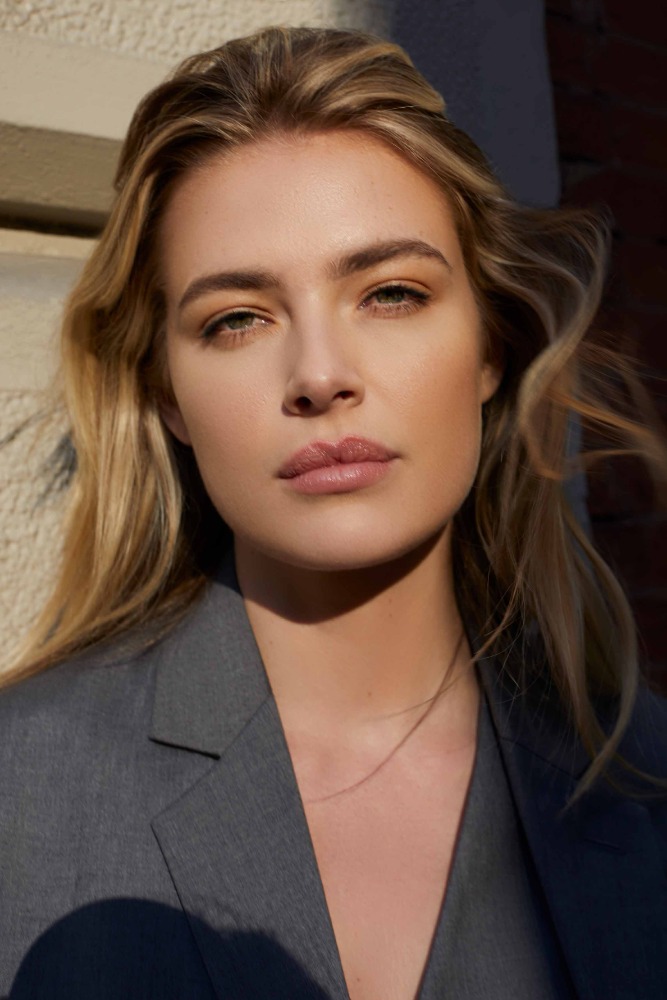 Close-up portrait of an adult woman with blonde hair wearing a gray blazer outdoors in natural light, looking directly at the camera.