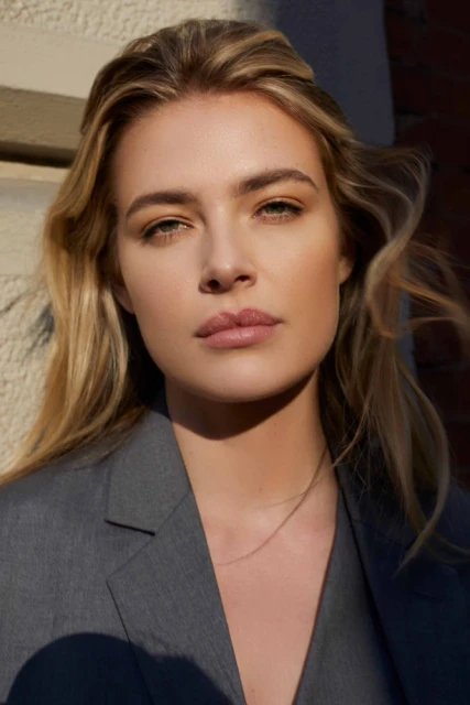 Close-up portrait of an adult woman with blonde hair wearing a gray blazer outdoors in natural light, looking directly at the camera.