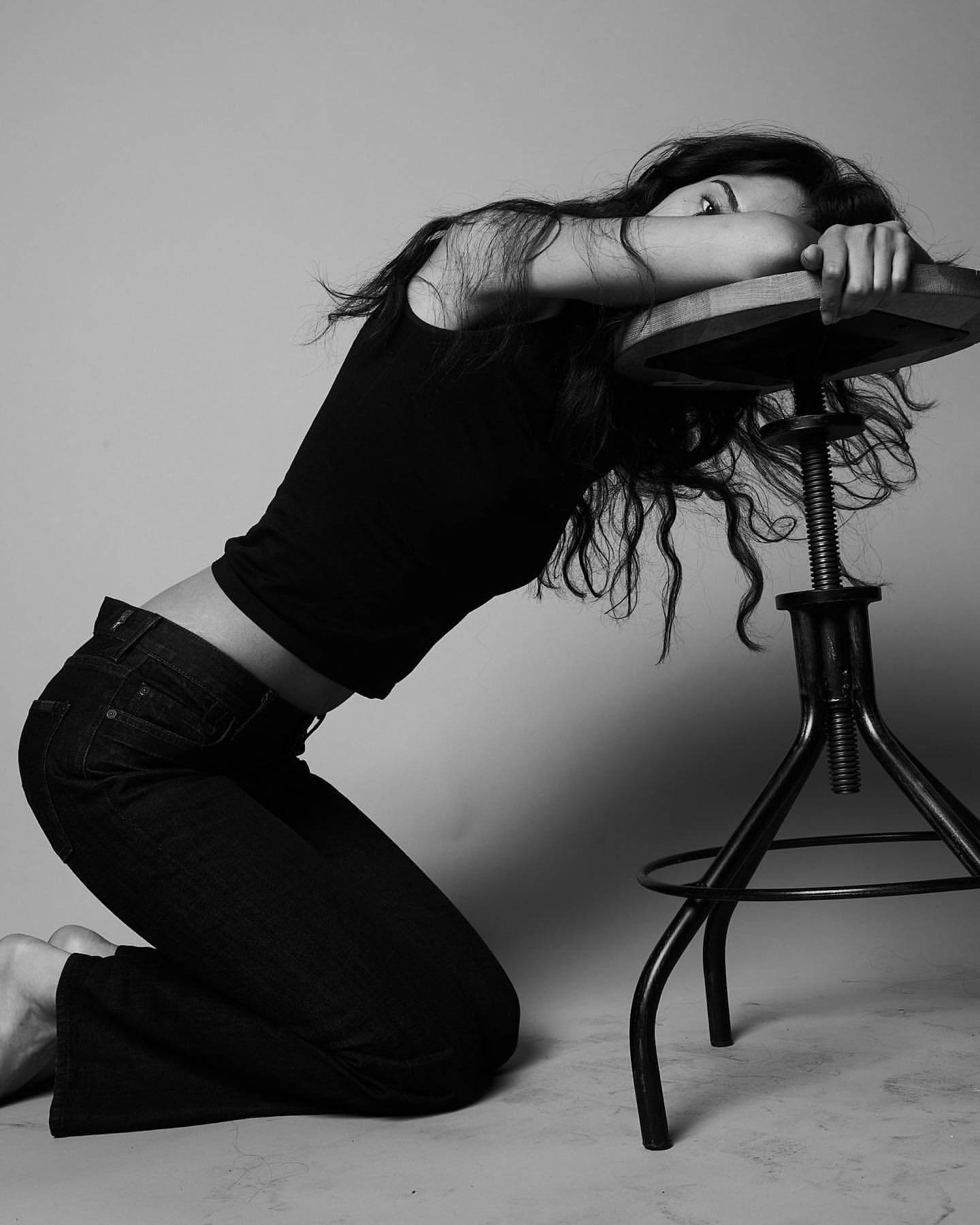 Monochrome portrait of a brunette woman kneeling with her head resting on a stool, wearing a black tank top and dark jeans.