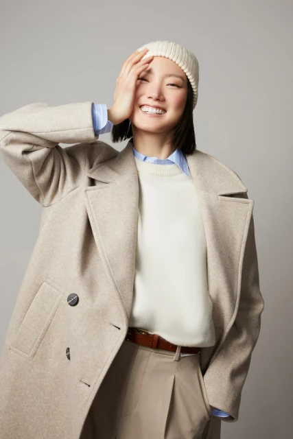 Portrait of an Asian woman smiling while wearing a winter hat, coat, sweater, and pants, against a neutral studio backdrop.